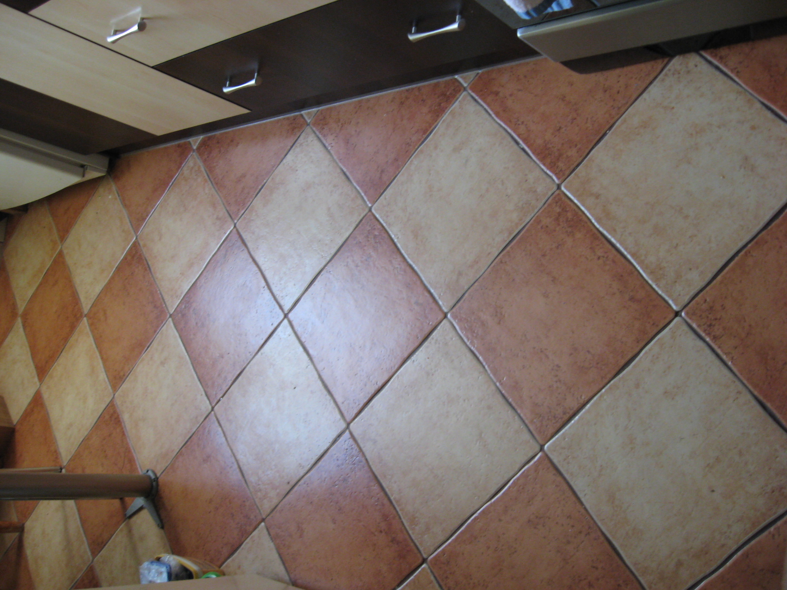 Ułożone ukośnie, kwadratowe płytki podłogowe w odcieniach beżu i brązu, widoczne fragmenty mebli kuchennych.